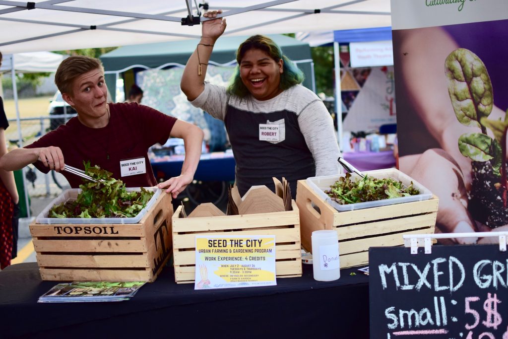 Two students smiling at their market booth, displaying their mixed greens.