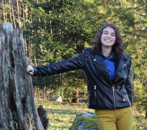 Valerie in front of some trees, leaning on a trunk, smiling