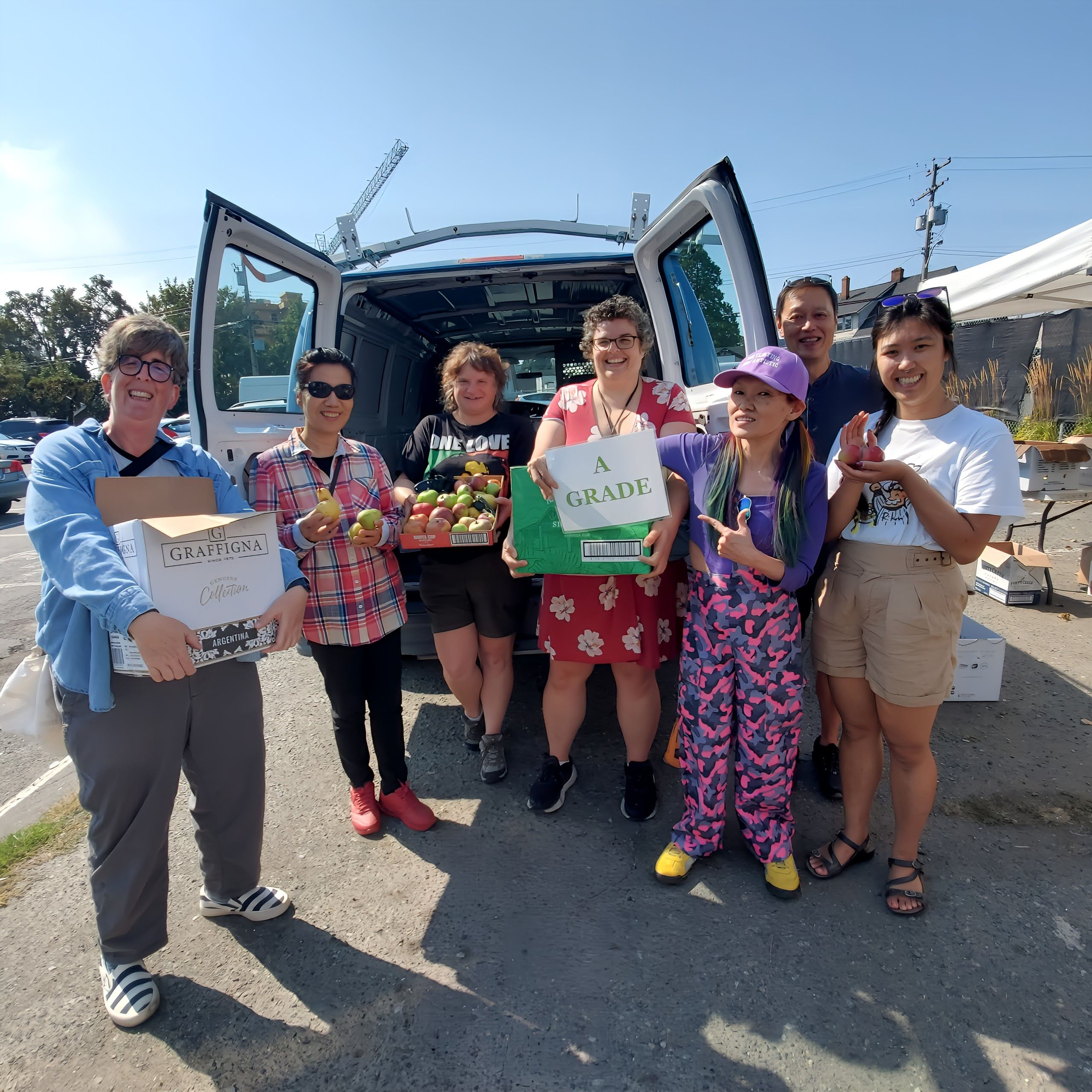 A group of six smiling volunteers standing in front of an open van, holding boxes of freshly picked apples labelled "A Grade".