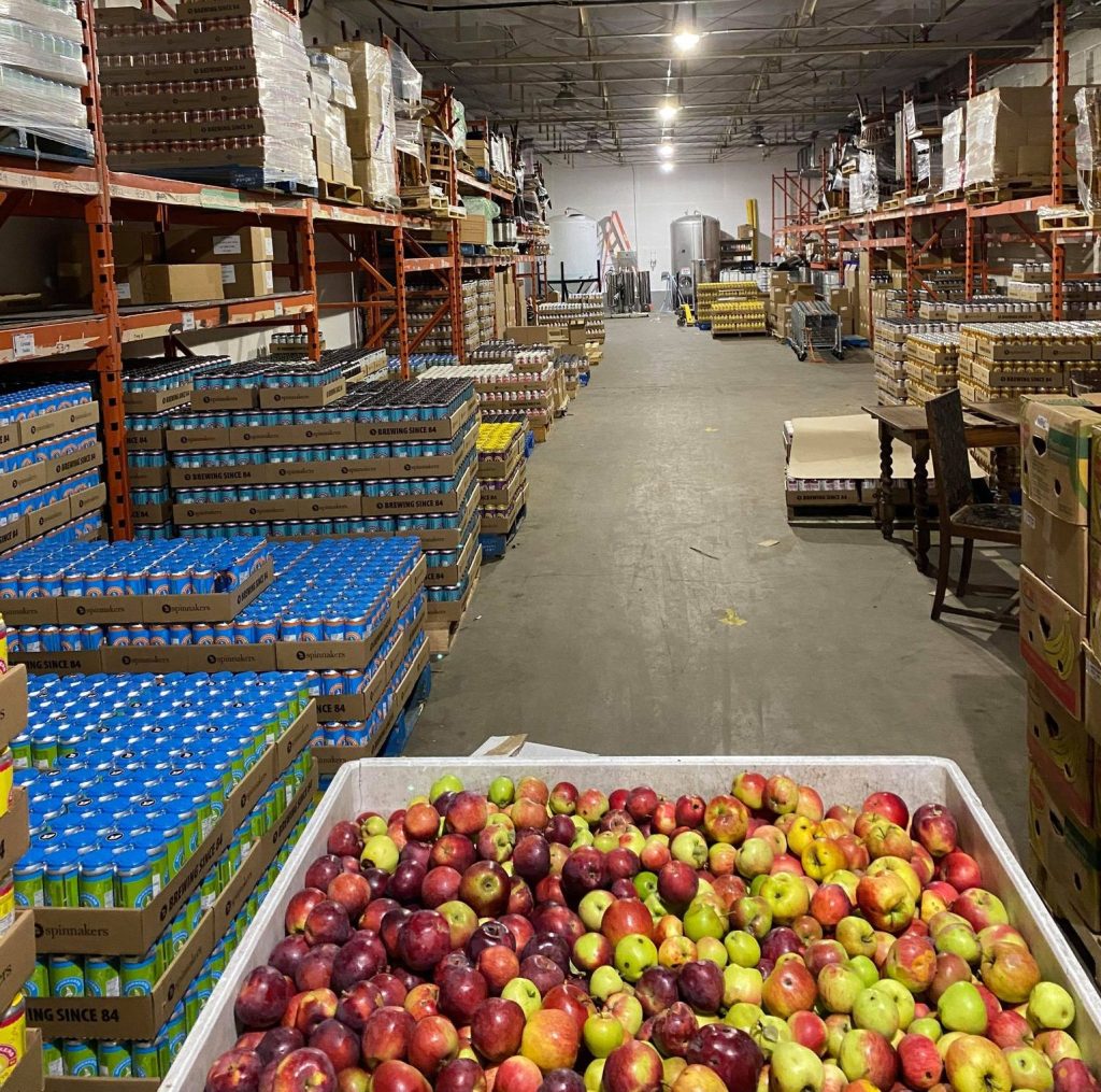 Lifecycles warehouse with a large bin of freshly picked red and green apples in the foreground, and stacks of boxed juice and cider on shelves behind.